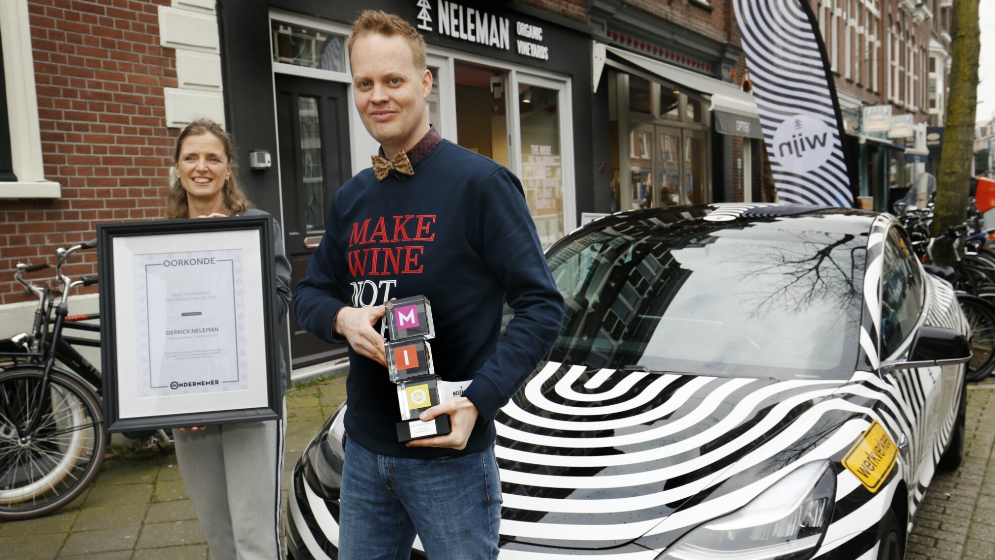 Wijnpionier Derrick Neleman en partner vorig jaar, met award en oorkonde, poserend voor hun zebra-Neleman-Tesla en brandstore in Utrecht. Foto: Ruud Voest.