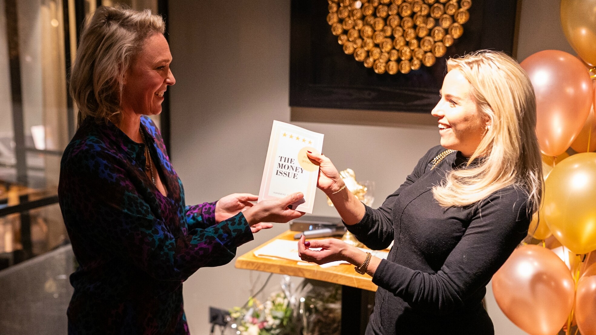Tijdens de drukbezochte boeklancering in Hotel Breezer in Amsterdam, overhandigde Hanneke van Onna (links) het eerste exemplaar van 'The Money Issue' aan Esther Vedder van het succesvolle sieradenmerk Vedder&amp;Vedder, 'voor mij, met haar tweelingzus Anne, een voorbeeld', aldus de auteur. Foto: Jeroen van der Wardt.