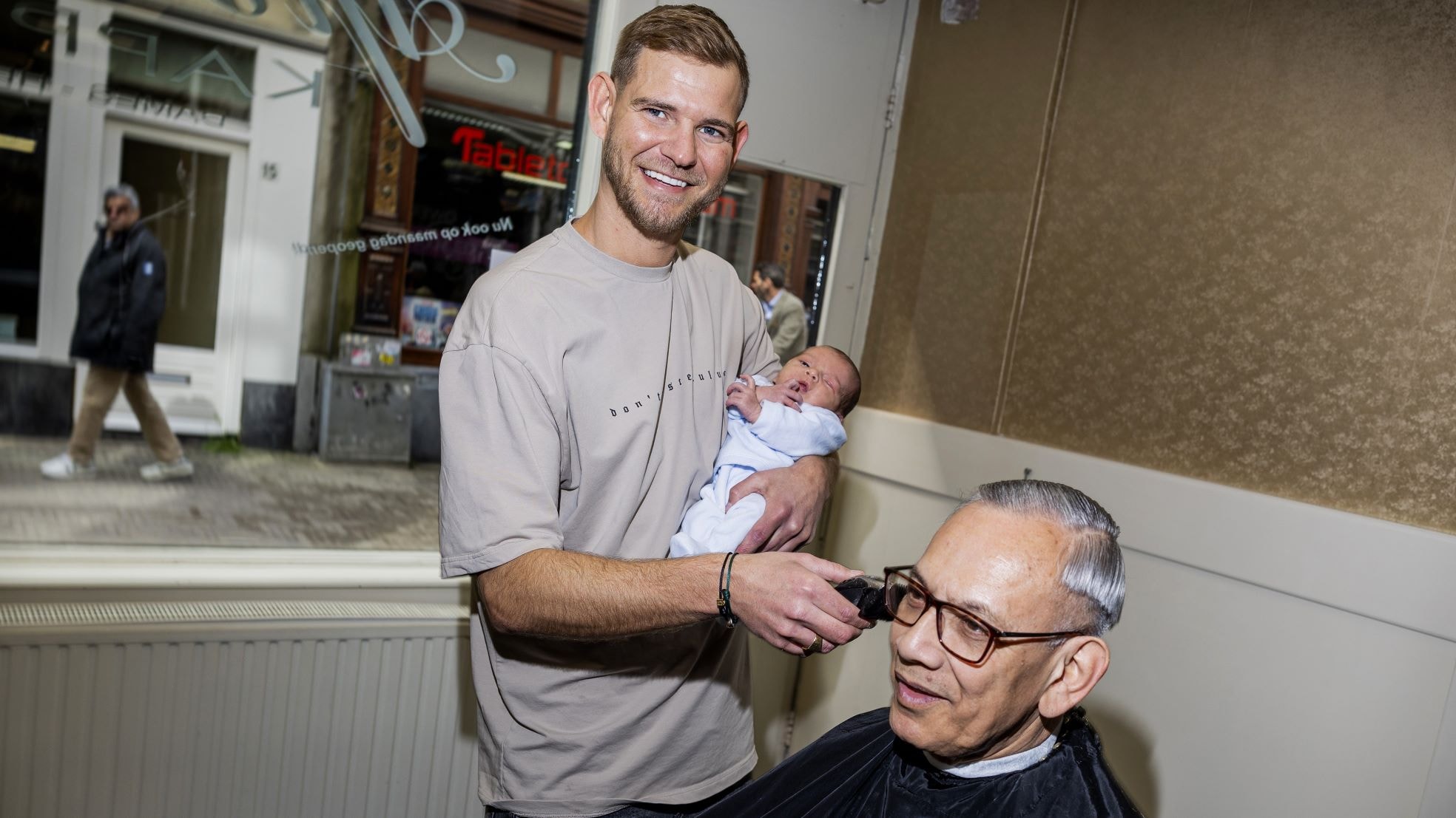 Nigel Israël scheert een klant met in zijn arm zijn twee weken jonge zoon Quincy. Foto: Guus Schoonewille Nigel Israël scheert een klant met in zijn arm zijn twee weken jonge zoon Quincy. Foto: Guus Schoonewille